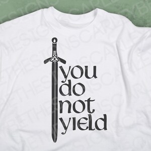 May include: White t-shirt with a black graphic of a sword and the words "you do not yield". The design is centered on the front of the shirt. The shirt is made of a soft material.