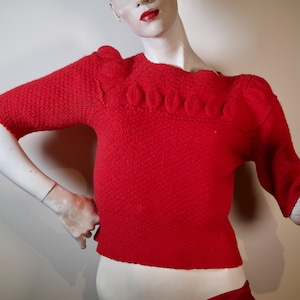 May include: A red knitted sweater with a textured pattern and a ruffled neckline. The sweater has short sleeves and a cropped length.