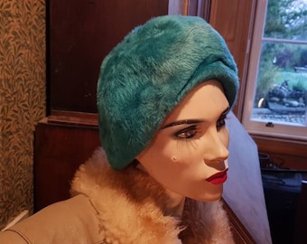 May include: A teal blue faux fur hat with a unique folded design. The hat is worn by a mannequin with a neutral expression.