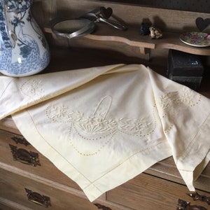 以下が含まれることがあります： 手縫いの花かごの刺繍が施された、繊細なクリーム色のリネン製のテーブルクロス。テーブルクロスは折り畳まれて、木製のドレッサーの上に置かれています。