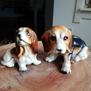 May include: Two ceramic figurines of brown and white dogs with black accents. The dogs are in a sitting position with their heads tilted up.