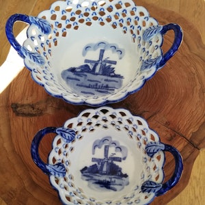 Peut inclure: Deux bols en céramique blanche avec des motifs de moulins à vent bleus et blancs. Les bols ont des côtés ajourés et des anses bleues.