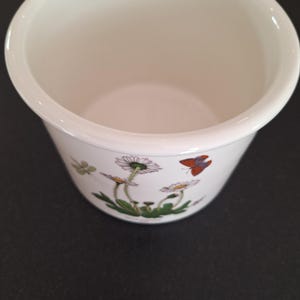 Pode incluir: Vaso de cerâmica branco com um design floral com margaritas brancas, folhas verdes e uma borboleta marrom.