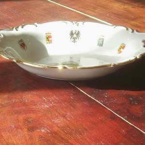 May include: White oval serving dish with gold trim and handles. The dish features several colourful crests and emblems. The rim has a decorative, scalloped edge. The dish is sitting on a wooden surface.