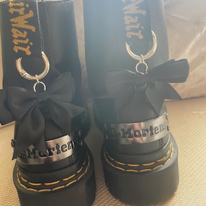May include: A pair of black leather boots with a platform sole and yellow stitching. Each boot has a black bow, a silver metal band with "Martens" and a yellow and black strap with "Dr. Martens".
