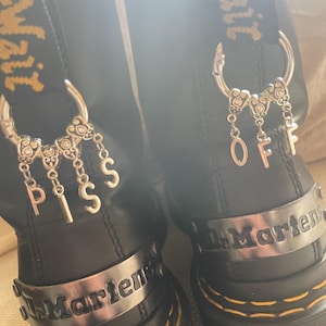 May include: Black leather boots with silver charms spelling "PISS" and "OFF". The boots have yellow stitching and a silver band with the brand name. The boots are a classic style.
