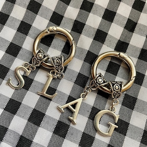 May include: Two silver keychains with letter charms. The charms spell out the word "SLAG".