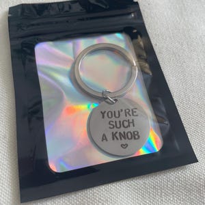 May include: A silver keyring with a circular charm, presented in a black plastic bag. The charm is engraved with the words "YOU'RE SUCH A KNOB" and a heart symbol. The bag has a holographic, iridescent backing.