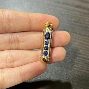 Könnte beinhalten: Goldener Anhänger mit zylindrischem Design, mit einer Reihe blauer Edelsteine. Der Anhänger ist mit kleinen, klaren Kristallen verziert und hat oben eine Schlaufe für eine Kette. Die Edelsteine haben unterschiedliche Formen und Größen.
