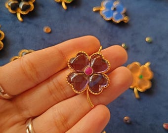 May include: A gold four-leaf clover pendant with a purple stone and a pink center. The pendant is held in a hand.