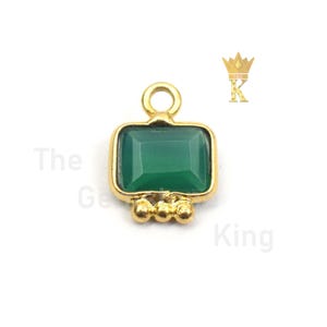 Pode incluir: Pingente dourado com uma gema verde engastada em uma moldura quadrada. O pingente tem um laço na parte superior para prender a uma corrente ou cordão. Há três pequenas contas douradas na parte inferior do pingente.