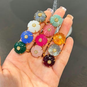 May include: A selection of gemstone rings in diverse colours, such as blue, green, pink, and orange. Each ring showcases a flower-shaped gemstone set in a gold band with a small diamond accent in the centre. The rings are displayed on a hand.