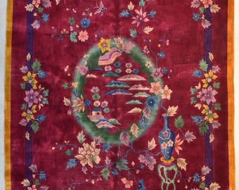 May include: A rectangular, burgundy-colored rug with a floral design. The rug features a central circular motif with a landscape scene and is framed by a border of flowers and leaves. The edges have a gold border and a dark blue geometric design.