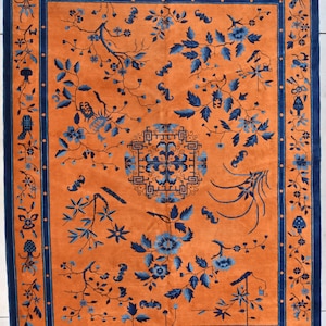 May include: A vintage Chinese rug with a blue and orange color scheme. The rug features a central design of a stylized flower with a geometric border. The rug is decorated with floral motifs and other traditional Chinese designs.