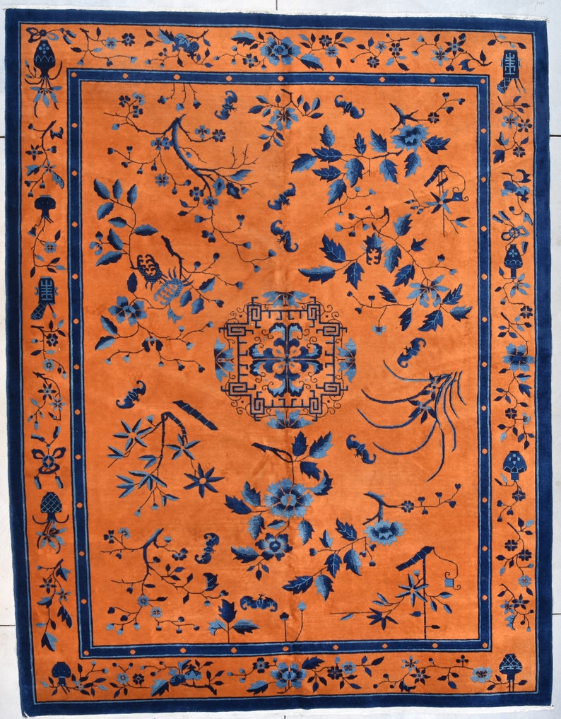 May include: A vintage Chinese rug with a blue and orange color scheme. The rug features a central design of a stylized flower with a geometric border. The rug is decorated with floral motifs and other traditional Chinese designs.