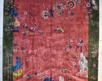 Op de afbeelding: Een vintage Chinees tapijt met een rode achtergrond en een groene rand. Het tapijt heeft een bloemenmotief met verschillende kleuren, waaronder roze, blauw en geel. Het tapijt is in goede staat en zou een mooie aanvulling zijn op elk huis.