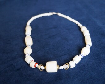 May include: A necklace featuring a series of white cylindrical and oval beads interspersed with small blue and white striped spacers, a single red ring, two gold-toned metal rings, and small clear beads, all strung together on a thin cord.