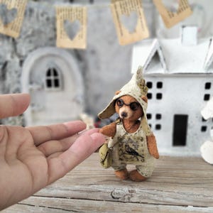 May include: A handcrafted teddy bear wearing a hat, glasses, and overalls with a bird design. The bear is holding a small bag and standing in front of a miniature house. Decorative paper hearts hang in the background.