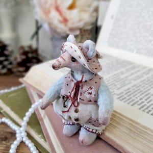 May include: A handmade, miniature mouse doll wearing a pink and white polka-dot hat and vest with a key pendant. The doll has blue arms and legs and is posed on top of an open book. A string of white pearls is in the foreground.