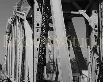 May include: A black and white photograph of a metal bridge with a truss design. The bridge is constructed from steel beams and has a series of vertical supports. The bridge is over a body of water, although the water is not visible in the image.