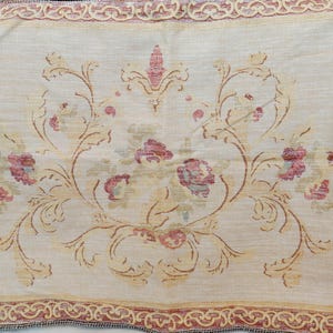 May include: A rectangular fabric placemat featuring a cream-colored base with a central floral motif in shades of muted red, pink, and sage green. The border consists of a repeating intricate scrollwork pattern in deep red and gold tones.