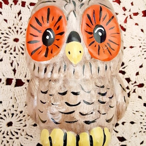 May include: A ceramic owl figurine with large orange eyes, a yellow beak, and brown and black painted feathers. The owl has yellow feet with black stripes.