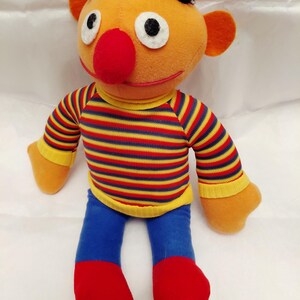 Puede incluir: Un peluche de un personaje con piel naranja, nariz roja y pelo negro. El personaje viste una camisa a rayas rojas, amarillas y azules, pantalones azules y zapatos rojos. El juguete tiene ojos blancos grandes y orejas naranjas.