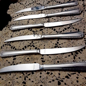 Könnte beinhalten: Sechs silberne Essmesser mit verzierten Griffen und gezahnten Klingen. Die Messer sind auf einer weißen Häkeldeckchen angeordnet.
