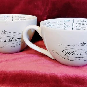 Puede incluir: Dos tazas de cerámica blanca con asa, cada una con el texto negro Café de Paris y un emblema de flor de lis en la parte frontal. Los bordes interiores están impresos con marcas de medición y nombres de bebidas como café filtre y lait chaud.
