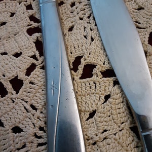 May include: Two silver flatware knives with a star design on the handle. The knives are on a white crocheted doily.