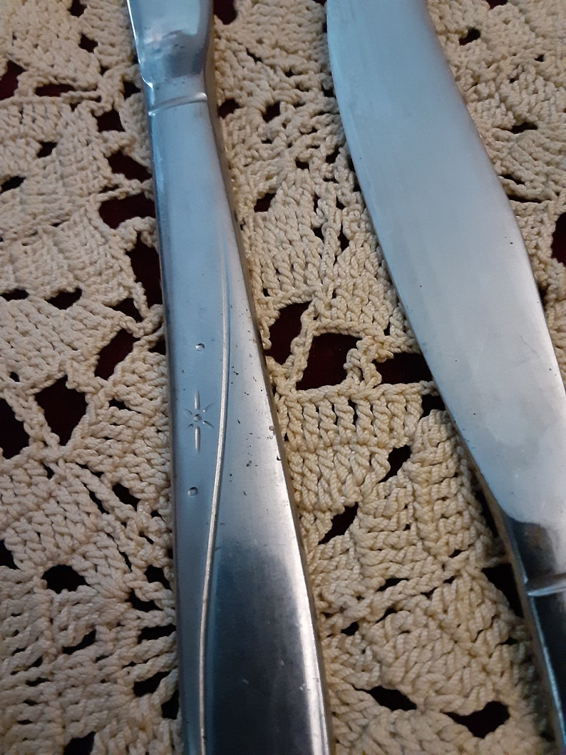 May include: Two silver flatware knives with a star design on the handle. The knives are on a white crocheted doily.
