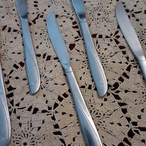 May include: Five silver dinner knives with a star design on the handle. The knives are laid out on a white crocheted doily.