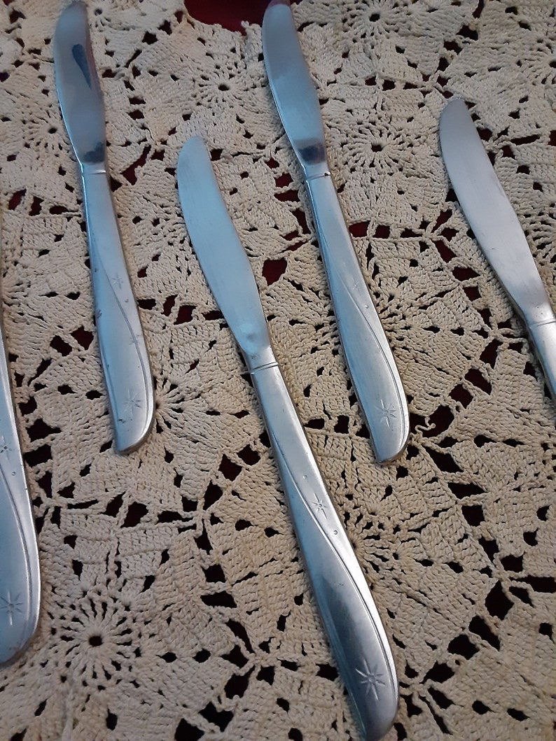 May include: Five silver dinner knives with a star design on the handle. The knives are laid out on a white crocheted doily.
