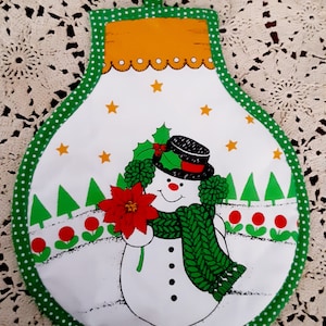 Puede incluir: Un agarrador de olla de Navidad verde y blanco con un muñeco de nieve sosteniendo una flor de pascua. El muñeco de nieve lleva un sombrero negro y una bufanda verde. El agarrador de olla tiene lunares blancos en el borde verde.