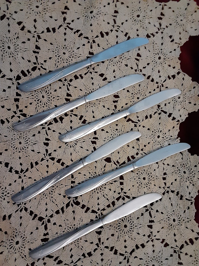 May include: Five silver dinner knives with a star design on a white crocheted tablecloth.