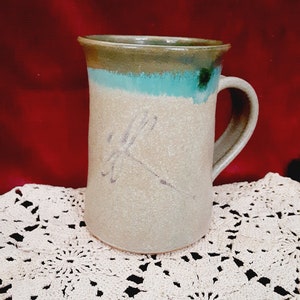 May include: A ceramic mug with a light brown glaze and a green and blue glaze band around the top. The mug has a simple design of a stylized flower on the front.