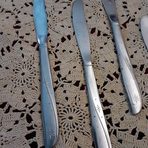 May include: Three silver dinner knives with star-shaped designs on a white crocheted doily.