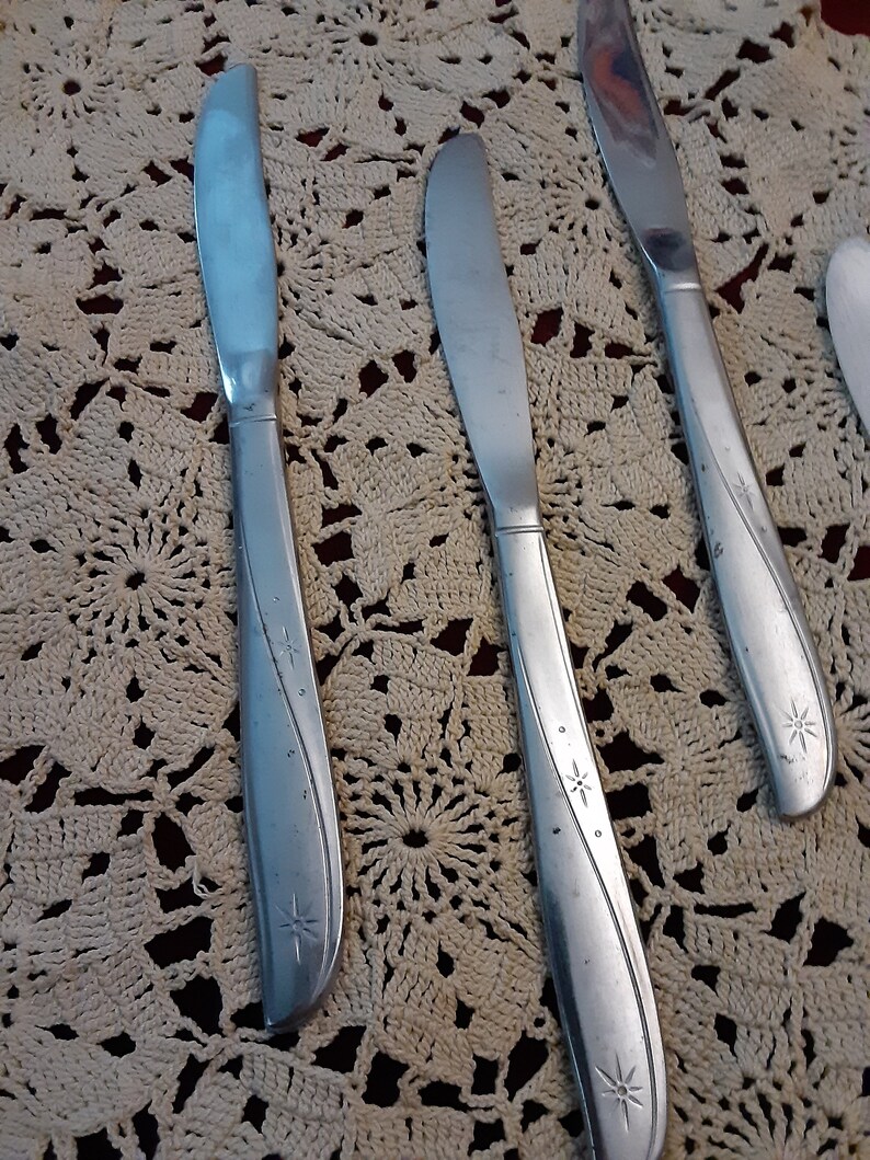 May include: Three silver dinner knives with star-shaped designs on a white crocheted doily.
