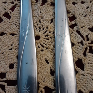 May include: Two silver flatware pieces with a star design on the handle. The flatware is on a crocheted surface.