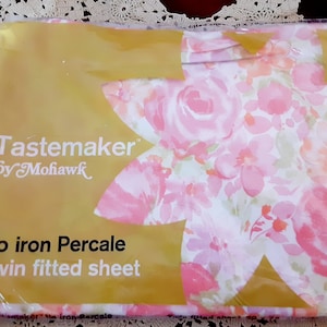 May include: A pink and white floral twin fitted sheet in a plastic package. The package has the text "Tastemaker by Mohawk" and "No iron Percale Twin fitted sheet".