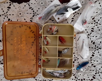 May include: A clear plastic tackle box with a brown lid that says "Magnum Custom Flies" on it. The box is open and contains a variety of fishing lures, including flies, spinners, and jigs. The lures are arranged in rows in the box. There are also several packages of fishing lures on the table next to the box.