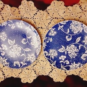 Könnte beinhalten: Zwei blaue und weiße Keramikteller mit Blumenmuster. Die Teller liegen auf einer gehäkelten Unterlage.