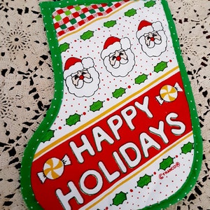 May include: A red and green Christmas stocking with a white background. It has three Santa faces, holly leaves, and candy canes. The text "HAPPY HOLIDAYS" is printed on the stocking. The stocking is trimmed with green and white polka dots.