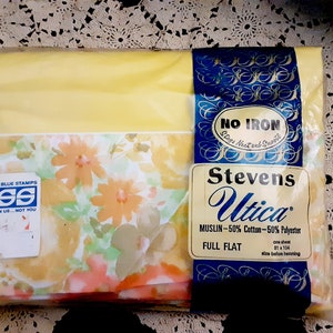 Puede incluir: Una sábana plana de flores amarillas y blancas en un paquete de plástico. El paquete tiene una etiqueta azul con el texto "Stevens Utica" y "Muslin - 50% algodón - 50% poliéster". La etiqueta también dice "Full Flat" y "una sábana 81 x 104 tamaño antes de doblar".