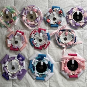 May include: A collection of ten circular rosette badges made from layered ribbons in various colors including pink, purple, blue, and white. Each badge features a central felt backing with a small black circular magnet and a white fabric loop.
