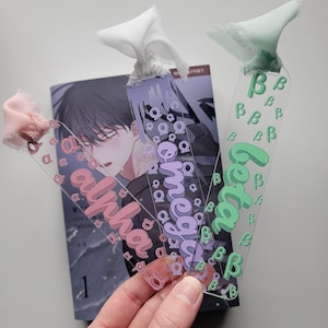 May include: Three clear acrylic bookmarks with different colored designs. The bookmarks feature the words "alpha", "omega", and "beta" in a stylized font. The bookmarks are decorated with repeating letters in a smaller font.