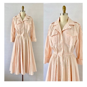 Pu&ograve; includere: Un abito vintage a righe rosa e bianche con chiusura a bottoni sul davanti, maniche lunghe e vita stretta. L'abito ha una gonna ampia e un colletto.