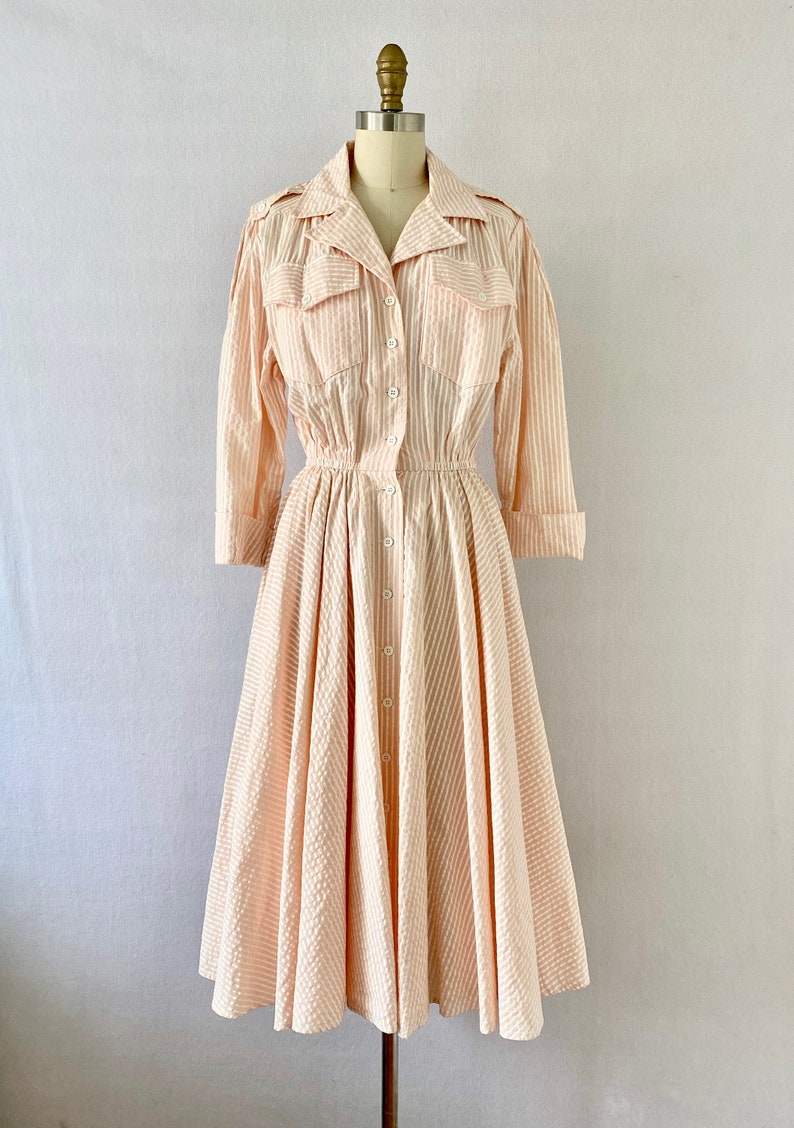 Pu&ograve; includere: Un abito vintage a righe rosa e bianche con chiusura a bottoni, maniche lunghe e gonna ampia. L'abito ha una vita stretta e una vestibilit&agrave; rilassata.