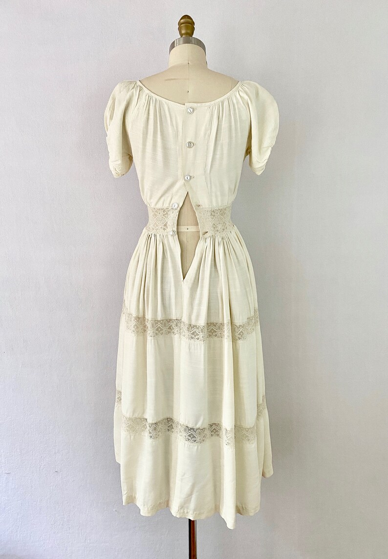 May include: A white vintage dress with a lace trim and a button-up back. The dress has a fitted bodice and a full skirt.