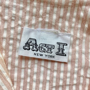 Pu&ograve; includere: Un'etichetta bianca con testo nero che recita "Act I NEW YORK" su un tessuto a righe rosa e bianche.